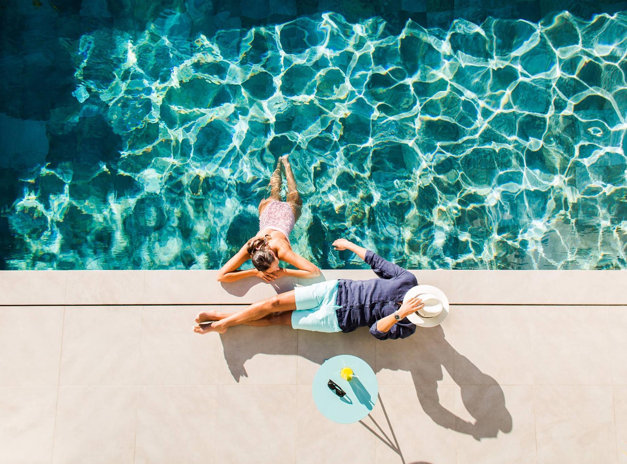 Overhead view of a woman in a blue swimming pool and a man on the sunny deck beside her.