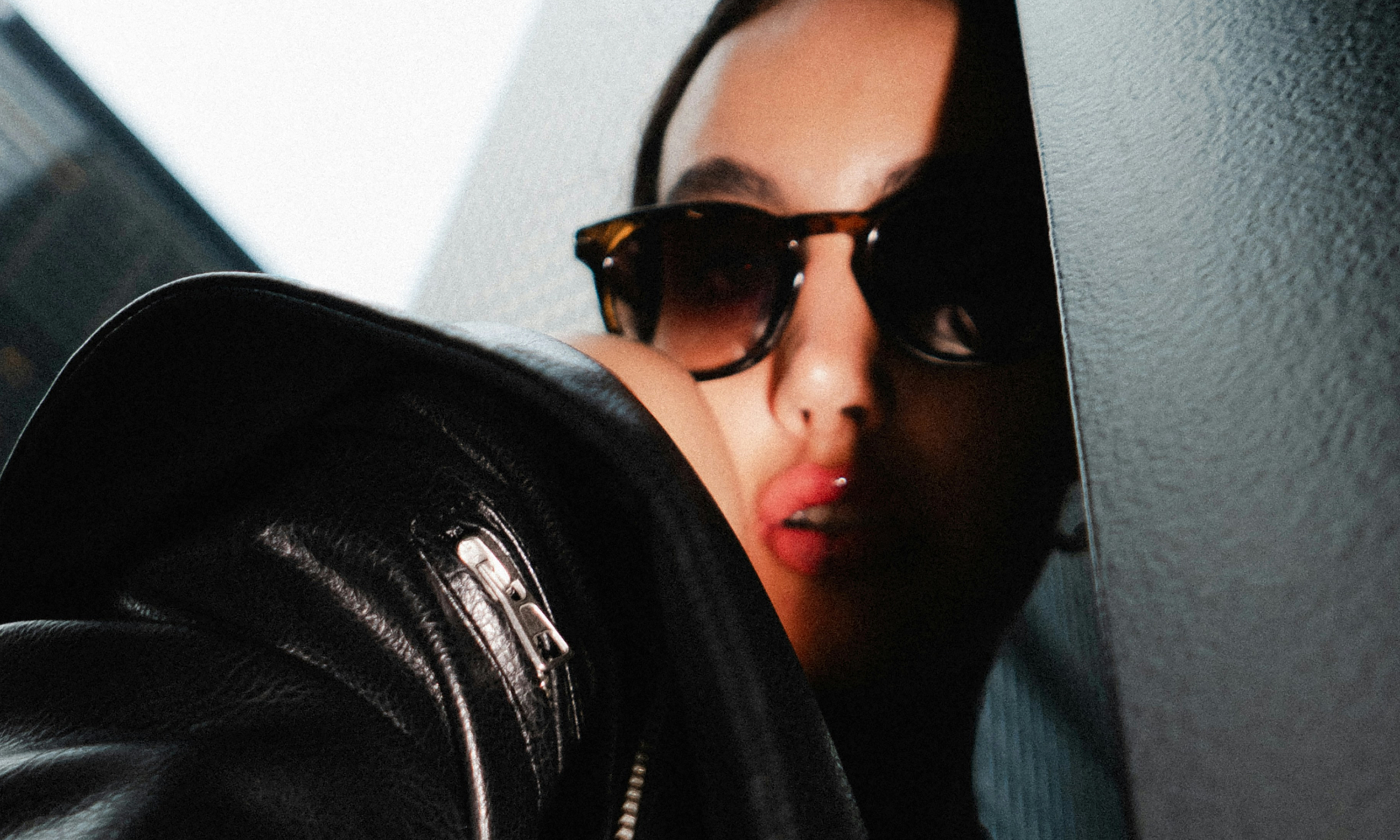 A woman in sunglasses and a black leather jacket with glossy lips, partially obscured by a dark wall.